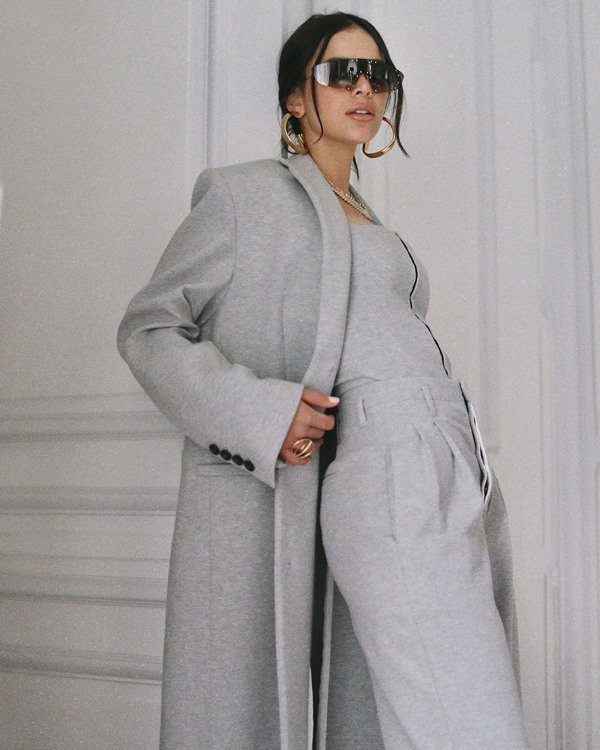 Actress Bruna Marquezin poses for a photo at the hotel door.  She is a young white woman with short straight hair, wearing a custom gray jacket, trousers, jacket and brown 2000s style sunglasses.